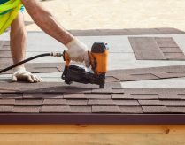 Construction worker putting the asphalt roofing (shingles) with nail gun on a new frame house