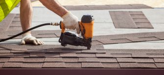 Construction worker putting the asphalt roofing (shingles) with nail gun on a new frame house