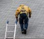 House during day with gray color Single Family Home and construction man in yellow uniform walking on roof shingles and ladder during repair