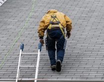 House during day with gray color Single Family Home and construction man in yellow uniform walking on roof shingles and ladder during repair