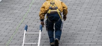 House during day with gray color Single Family Home and construction man in yellow uniform walking on roof shingles and ladder during repair