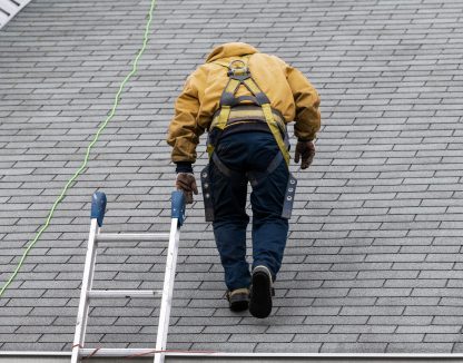 House during day with gray color Single Family Home and construction man in yellow uniform walking on roof shingles and ladder during repair