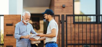 Happy mature man receiving package from a courier and signing paperwork in front of his house.