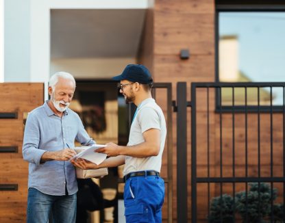 Happy mature man receiving package from a courier and signing paperwork in front of his house.