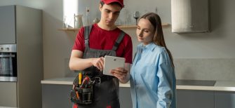 A professional technician, equipped with a tool belt, is consulting with a homeowner in a modern kitchen. The homeowner listens carefully as the technician explains the checklist. Both individuals are engaged in a discussion about the maintenance work that is either being proposed or has been completed.