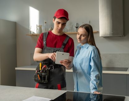 A professional technician, equipped with a tool belt, is consulting with a homeowner in a modern kitchen. The homeowner listens carefully as the technician explains the checklist. Both individuals are engaged in a discussion about the maintenance work that is either being proposed or has been completed.