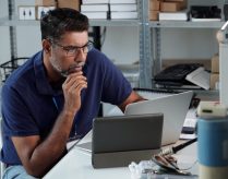 Man seated at desk, focusing on work with laptop and tablet present on desk Photographs and office supplies are arranged on shelves in background