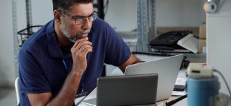 Man seated at desk, focusing on work with laptop and tablet present on desk Photographs and office supplies are arranged on shelves in background