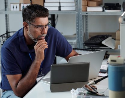 Man seated at desk, focusing on work with laptop and tablet present on desk Photographs and office supplies are arranged on shelves in background
